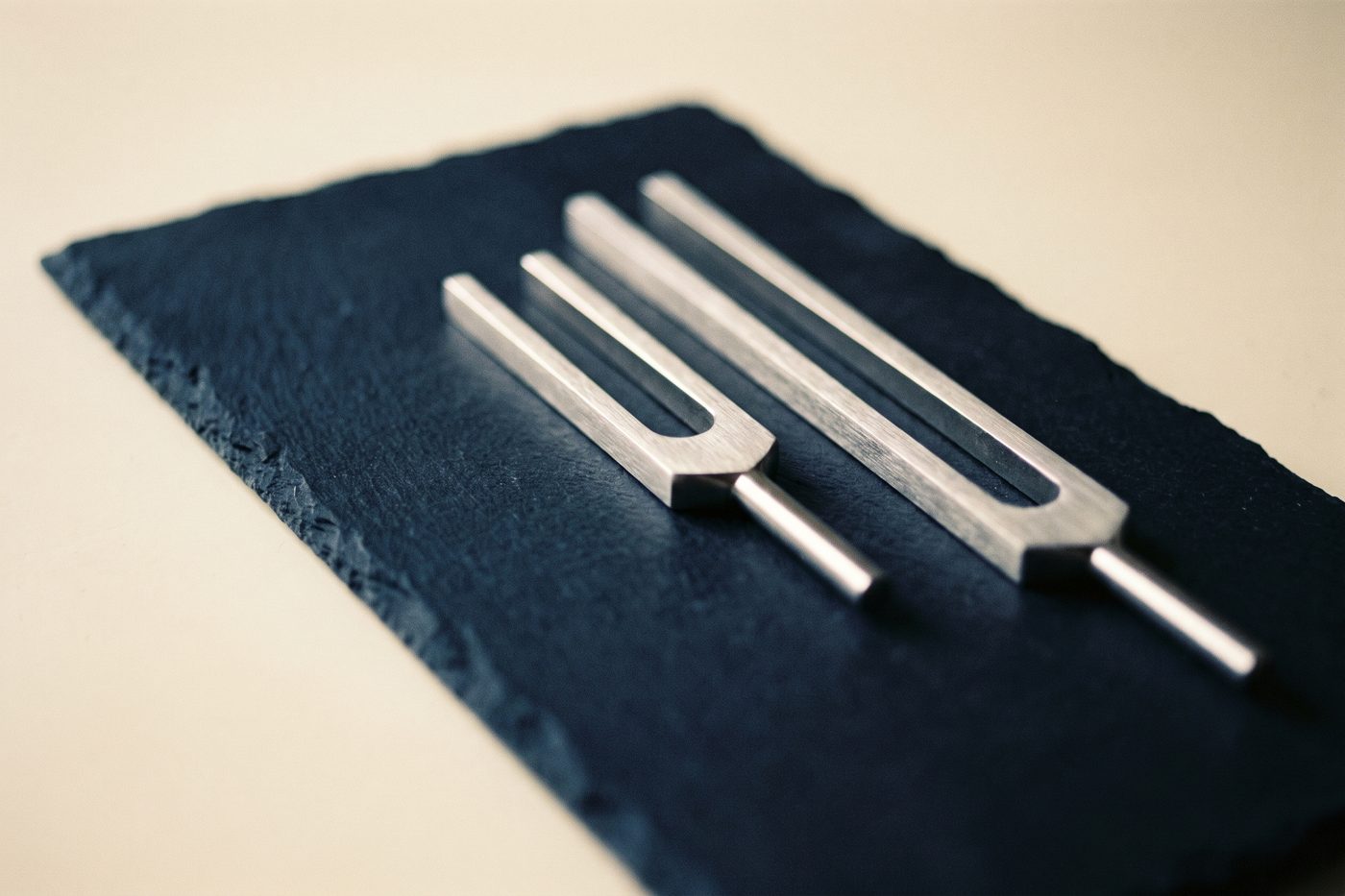Two brushed steel tuning forks of clearly different lengths side by side on navy slate, raking cream light