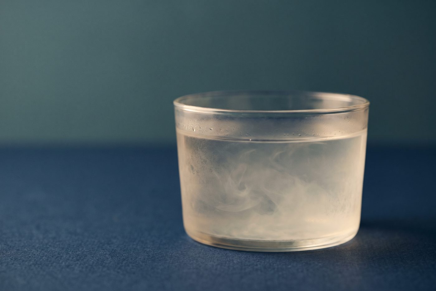 A glass vessel of clear liquid gone faintly cloudy, a soft haze suspended inside it, backlit teal on navy