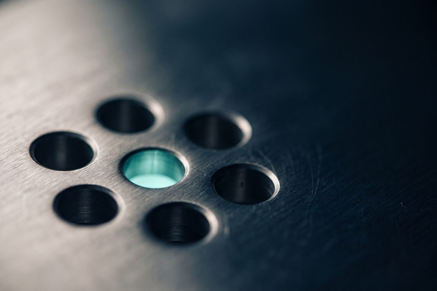 A cluster of machined circular ports in a brushed steel plate, one of them glowing teal from behind, extreme macro, navy