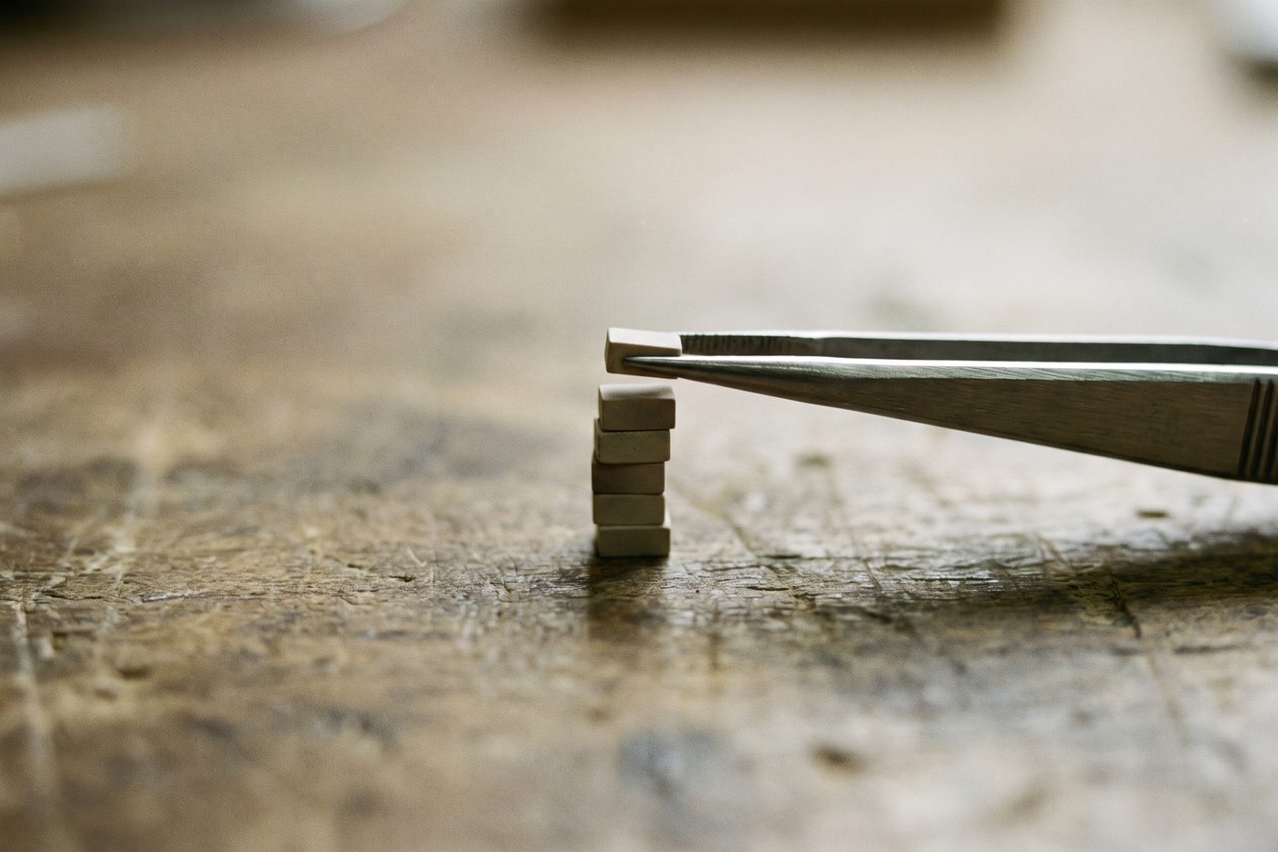 Tiny ceramic blocks being stacked one at a time into a precise column on a matte navy workbench