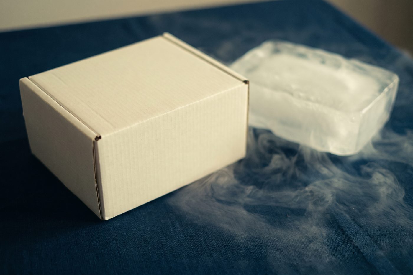 A plain unbranded cream corrugated box on a navy surface with a block of frosted ice beside it, cold vapour drifting across