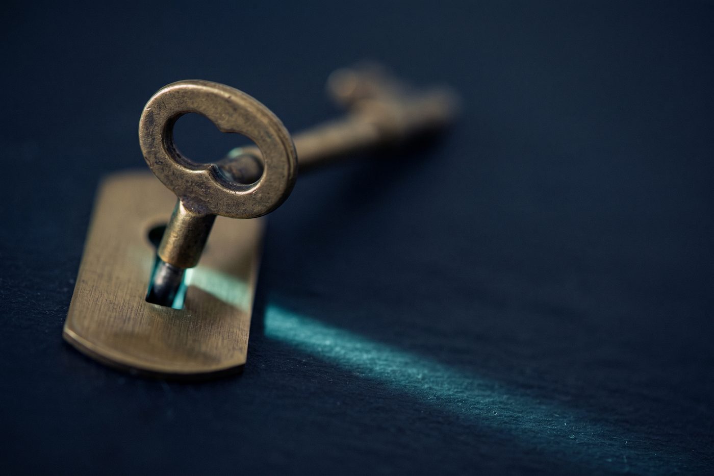 A brass key resting in a precisely machined keyhole plate on navy stone, teal light spilling through the slot