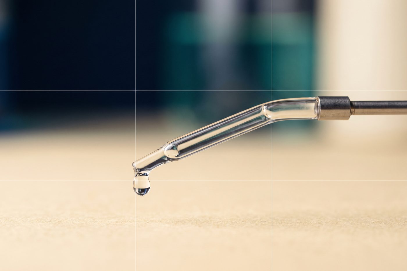 A glass column and fine steel tubing in extreme macro, a single clear droplet forming at the outlet, navy and teal