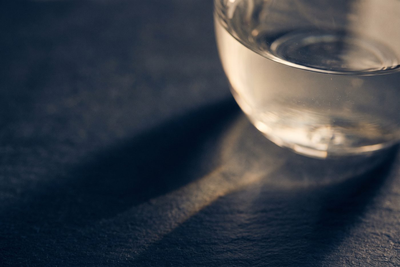 A glass vessel of clear liquid on navy slate casting a long sundial shadow across the surface, warm cream light