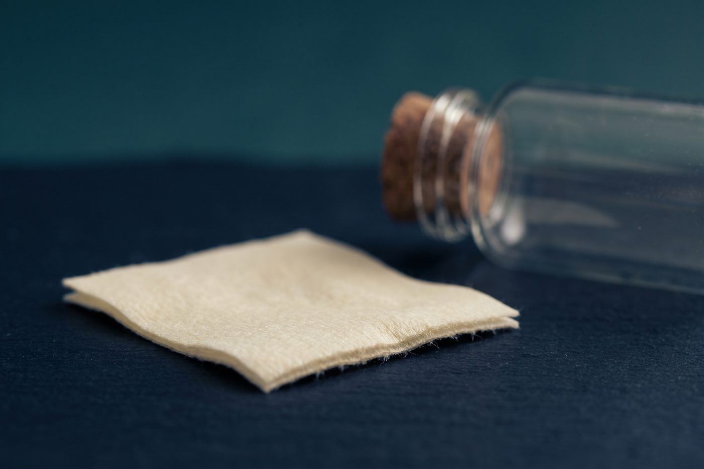 A single alcohol swab unfolded on matte navy stone beside a sealed glass vessel, cool light, sterile stillness