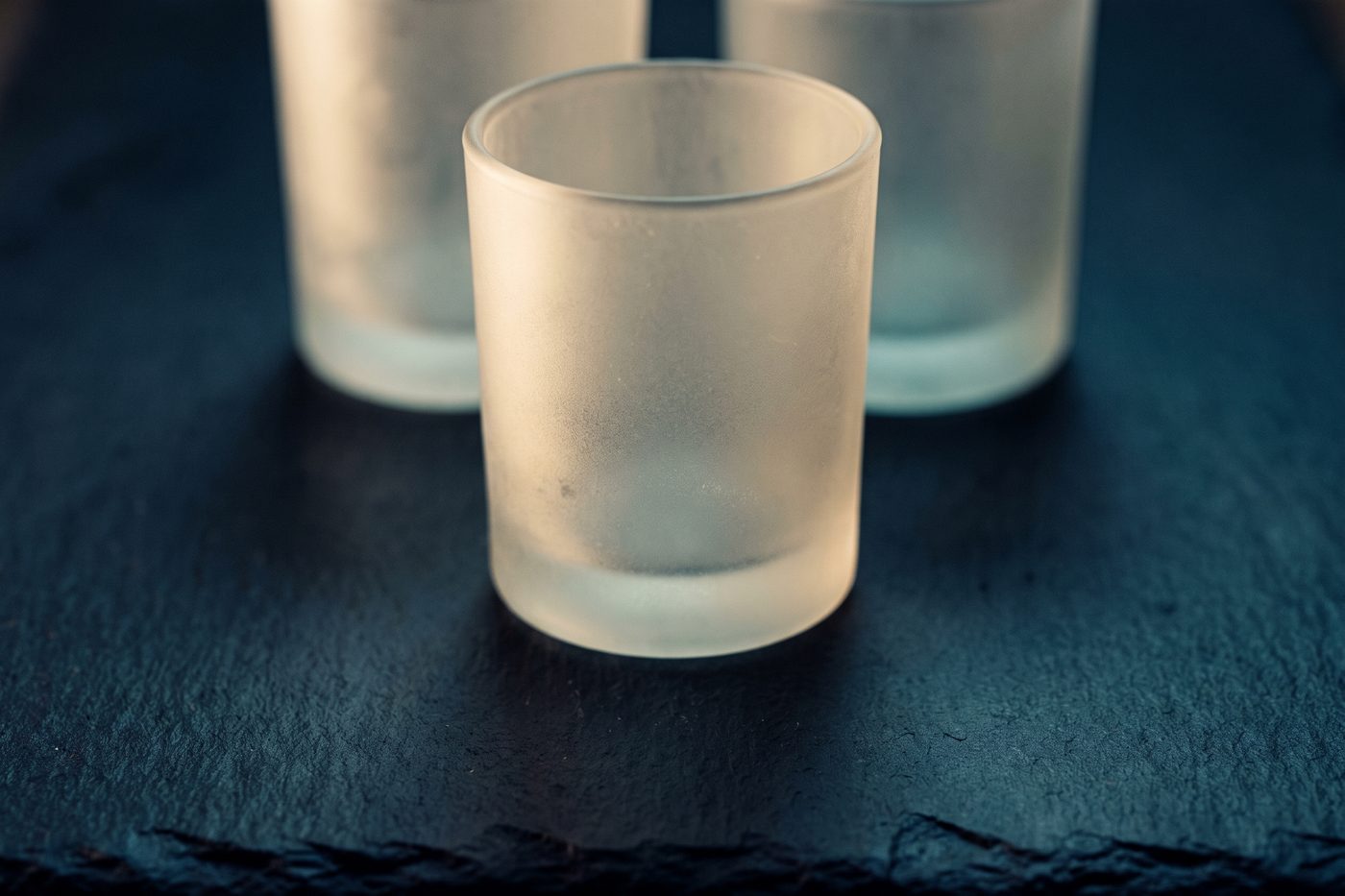 Three identical frosted glass vessels on navy slate, one lifted slightly forward into a pool of warm cream light