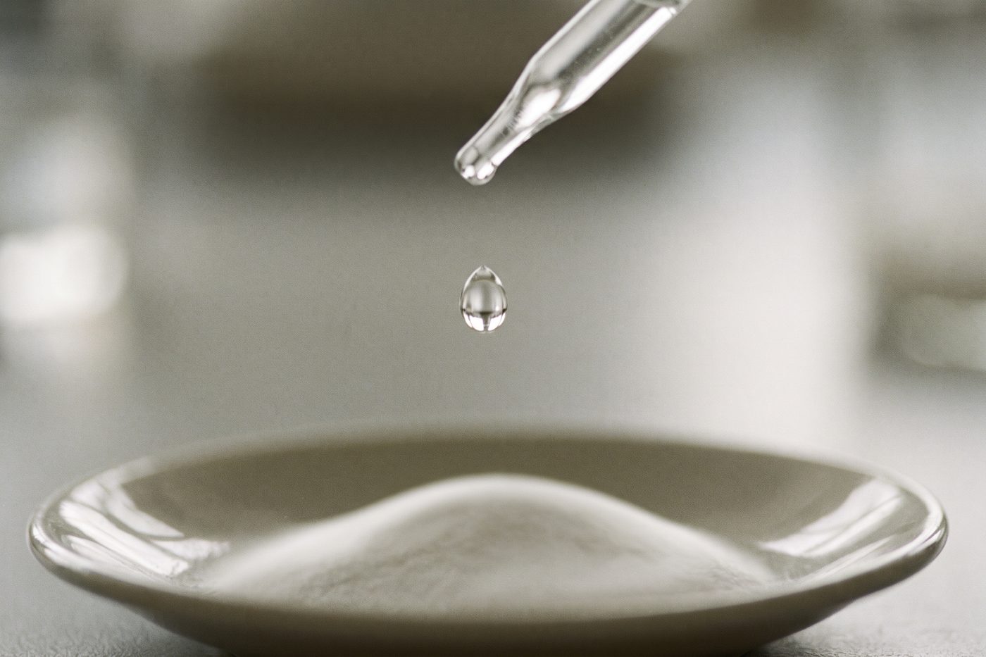 A clear droplet falling from a fine glass pipette toward a shallow dish of white powder, extreme macro, backlit teal against navy
