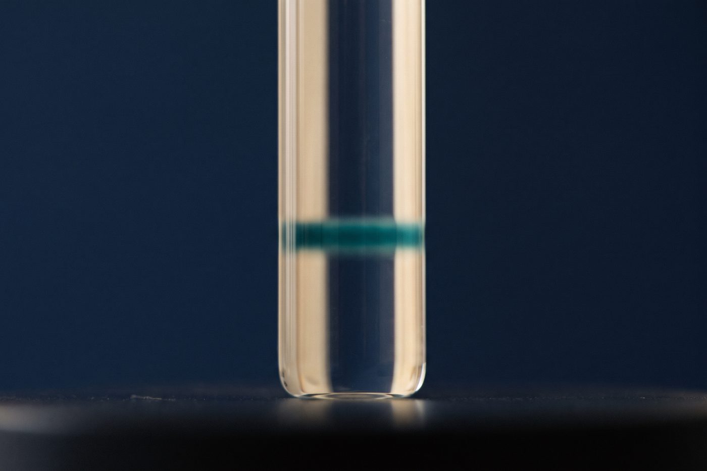 A clear glass chromatography column with a single sharp band of teal travelling down it, extreme macro, navy backdrop