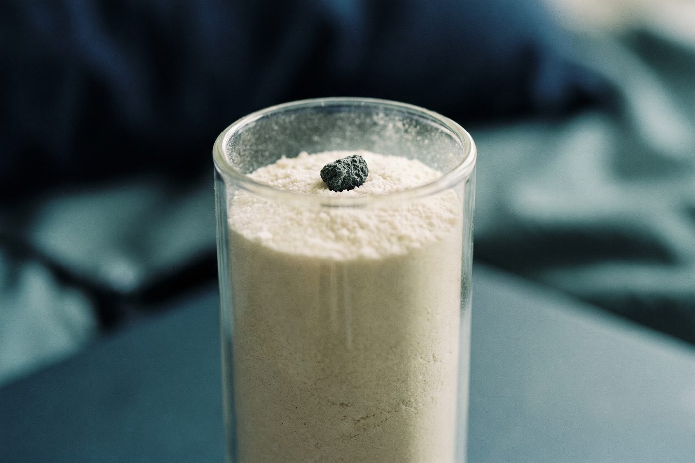 A tall glass cylinder of pale crystalline powder with one single dark grain visible near the top, extreme macro, navy