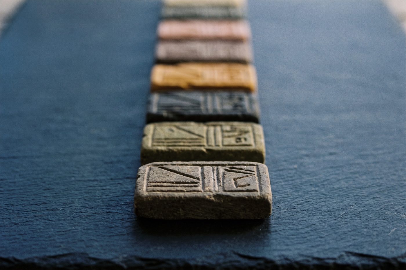 A row of small engraved stone tiles laid in a precise line on navy slate, each tile a slightly different muted tone
