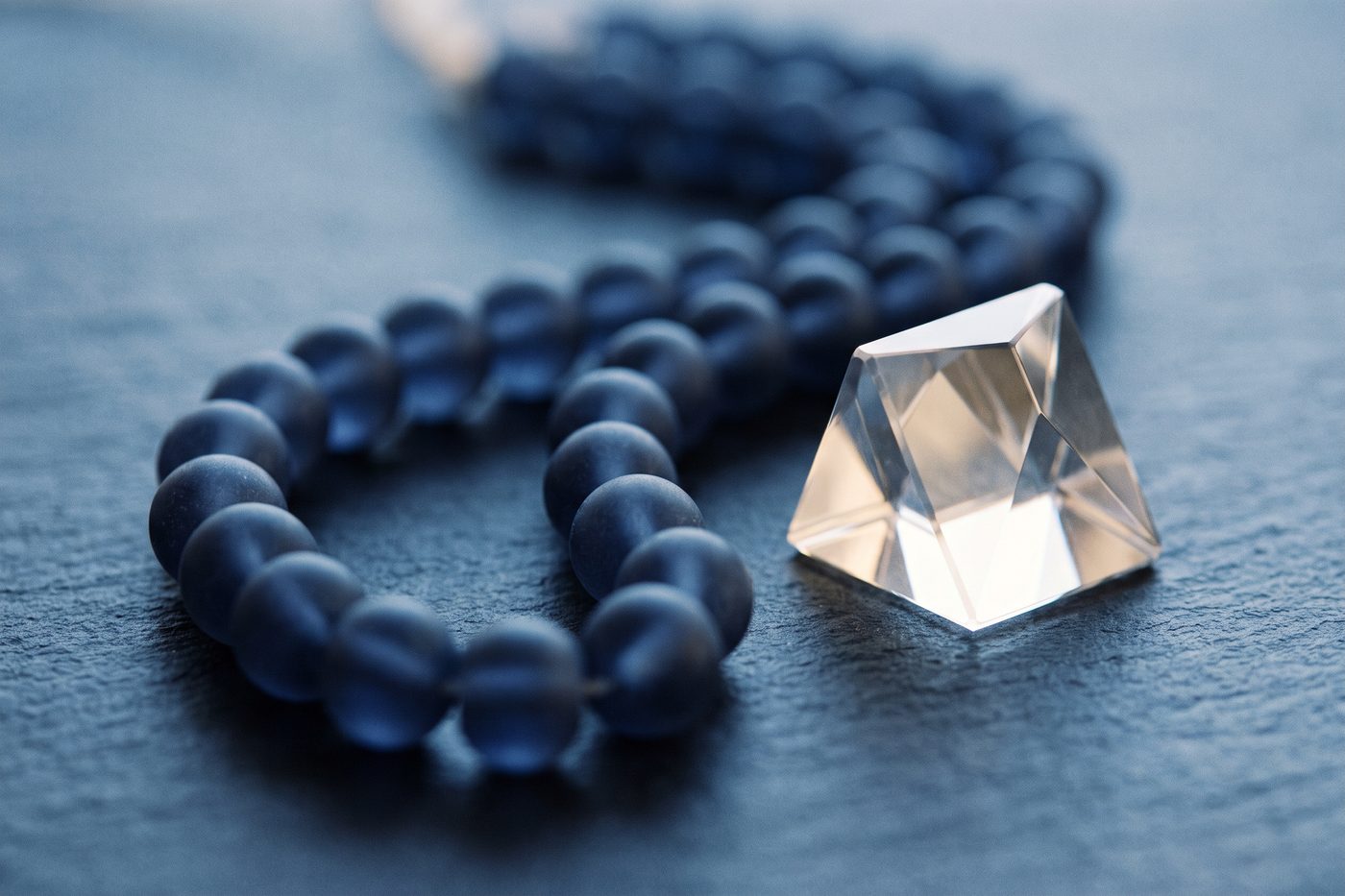 A chain of frosted glass beads beside a single hard cut crystal on matte navy slate, cool symmetrical light