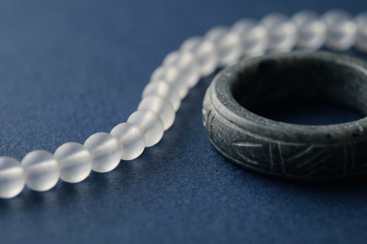 A fine chain of frosted beads beside a solid carved ring of dark stone on navy, raking light