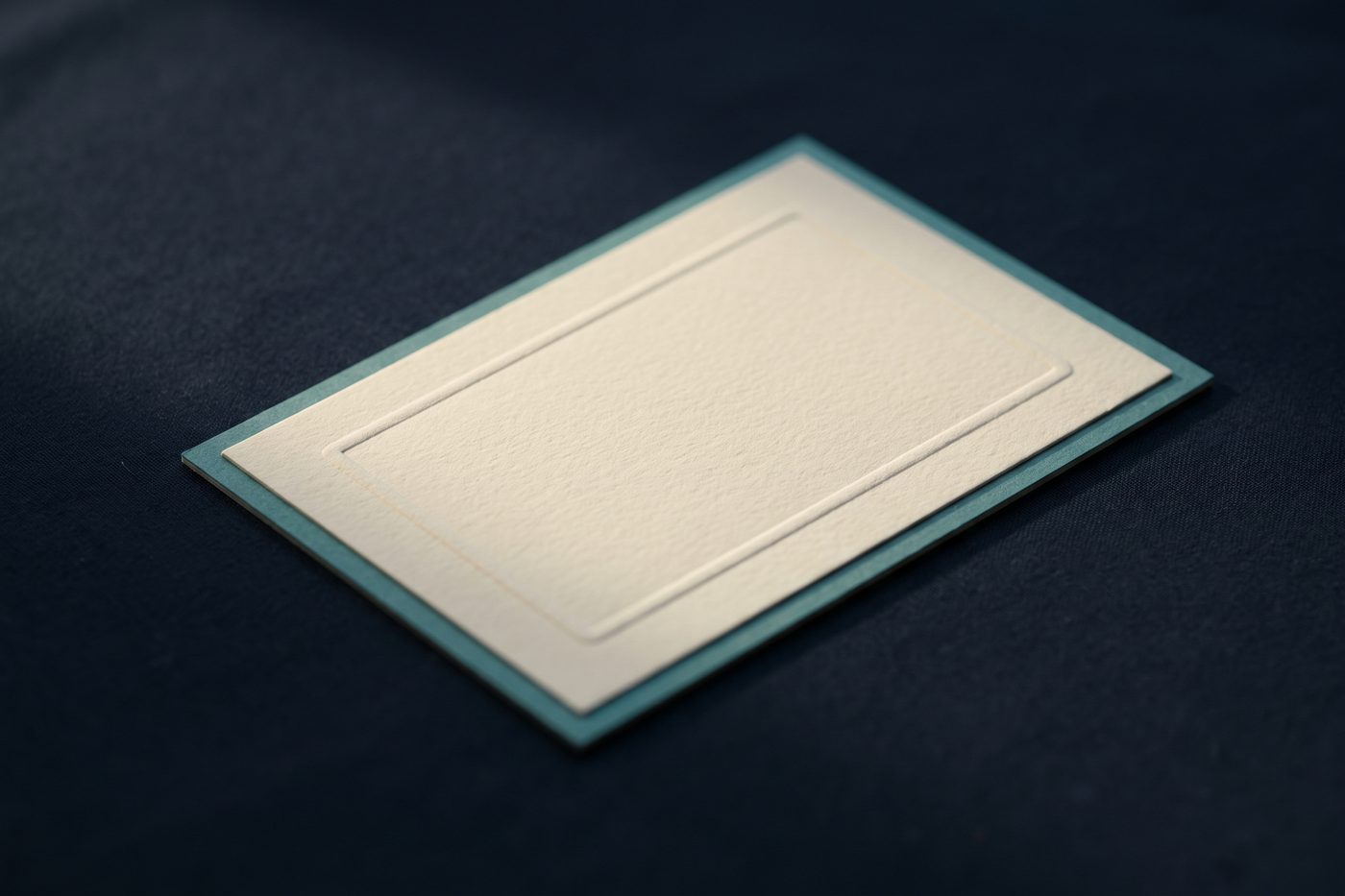 A blank embossed cream card with a thin teal border resting on matte navy, raking light picking up the deboss