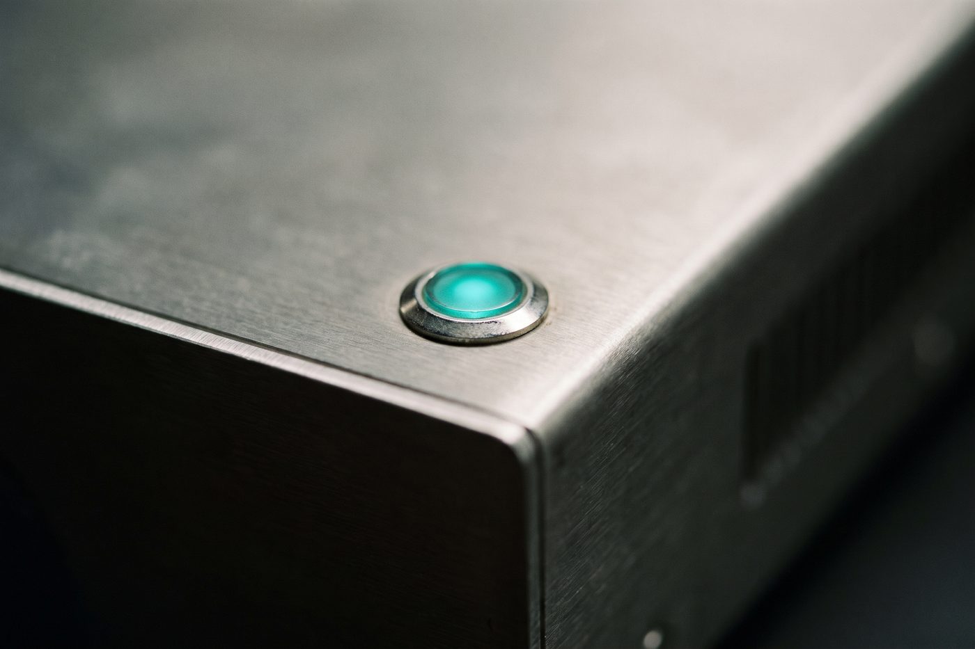 The brushed steel housing of an analytical instrument with a single small teal indicator light, extreme macro, shallow focus
