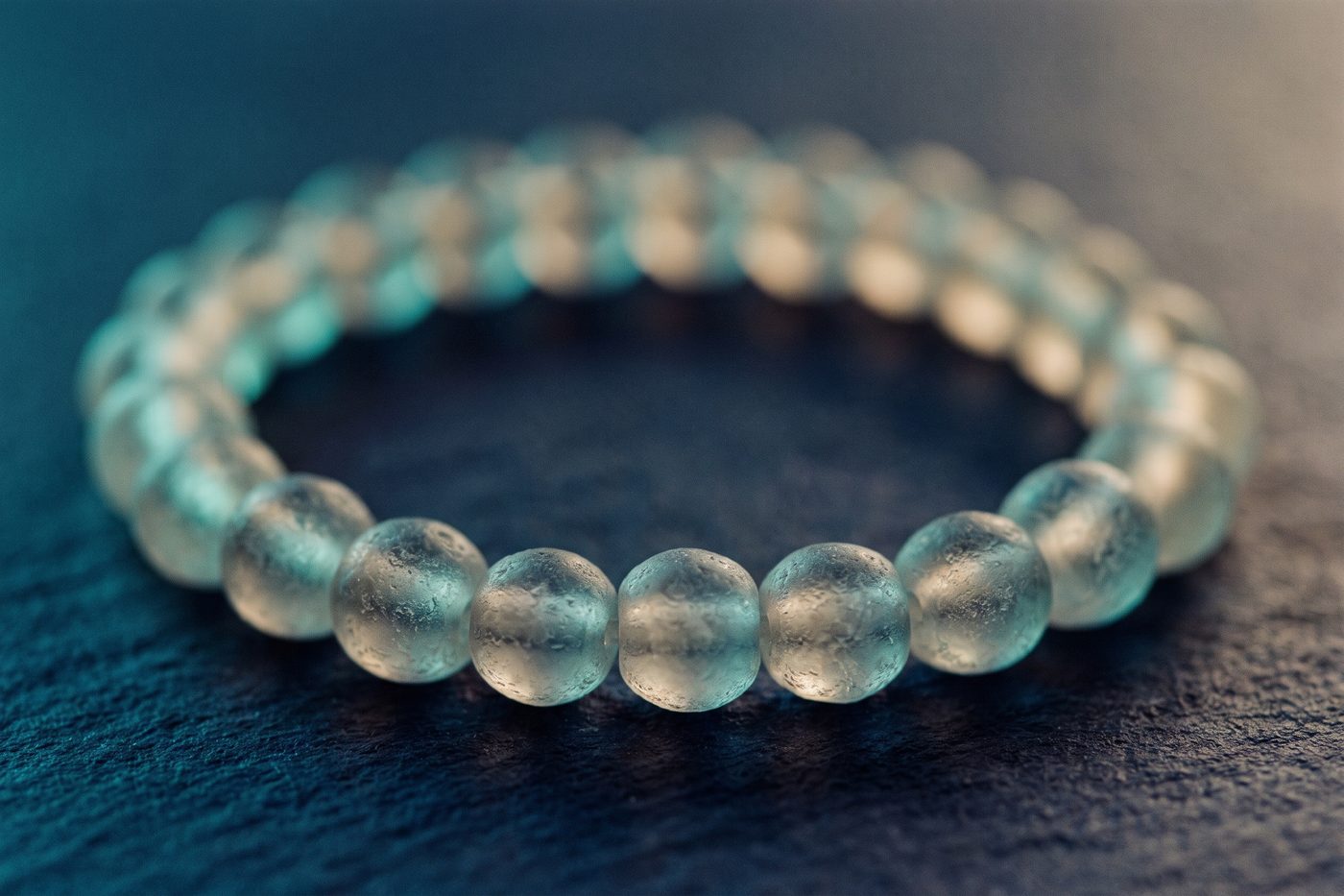 A closed ring of frosted glass beads forming a perfect circle on matte navy stone, rim lit in teal