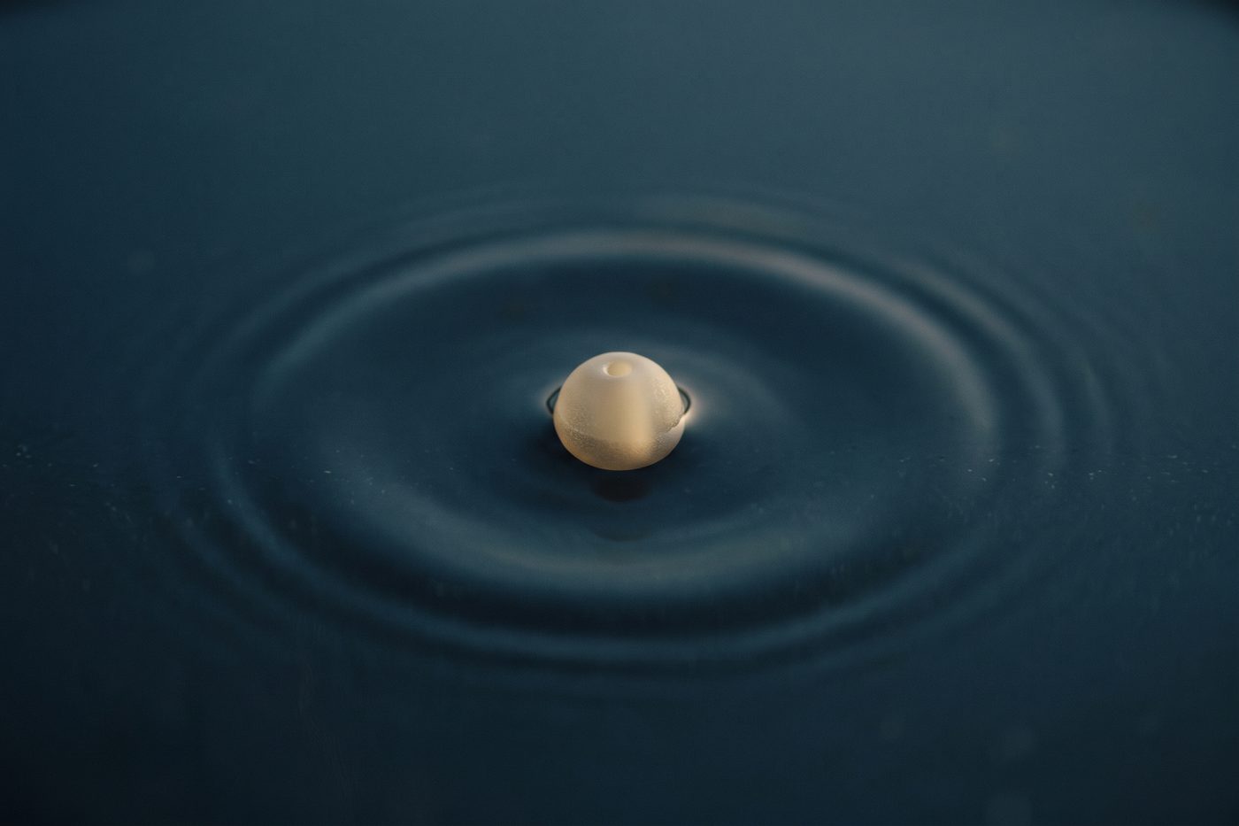 A single frosted glass bead suspended in still dark water, one faint ripple radiating outward, lit from above with cool teal light