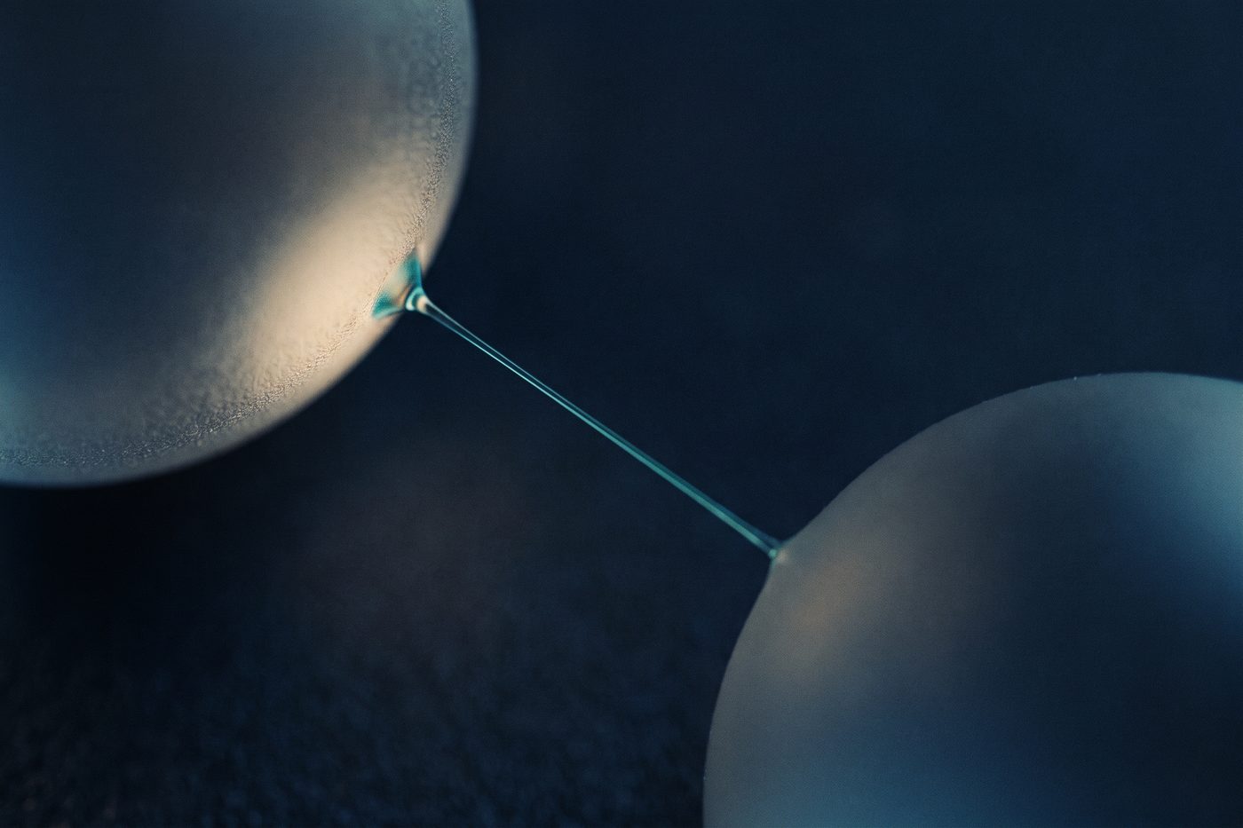 Two frosted glass spheres joined by a single fine glowing teal filament, extreme macro