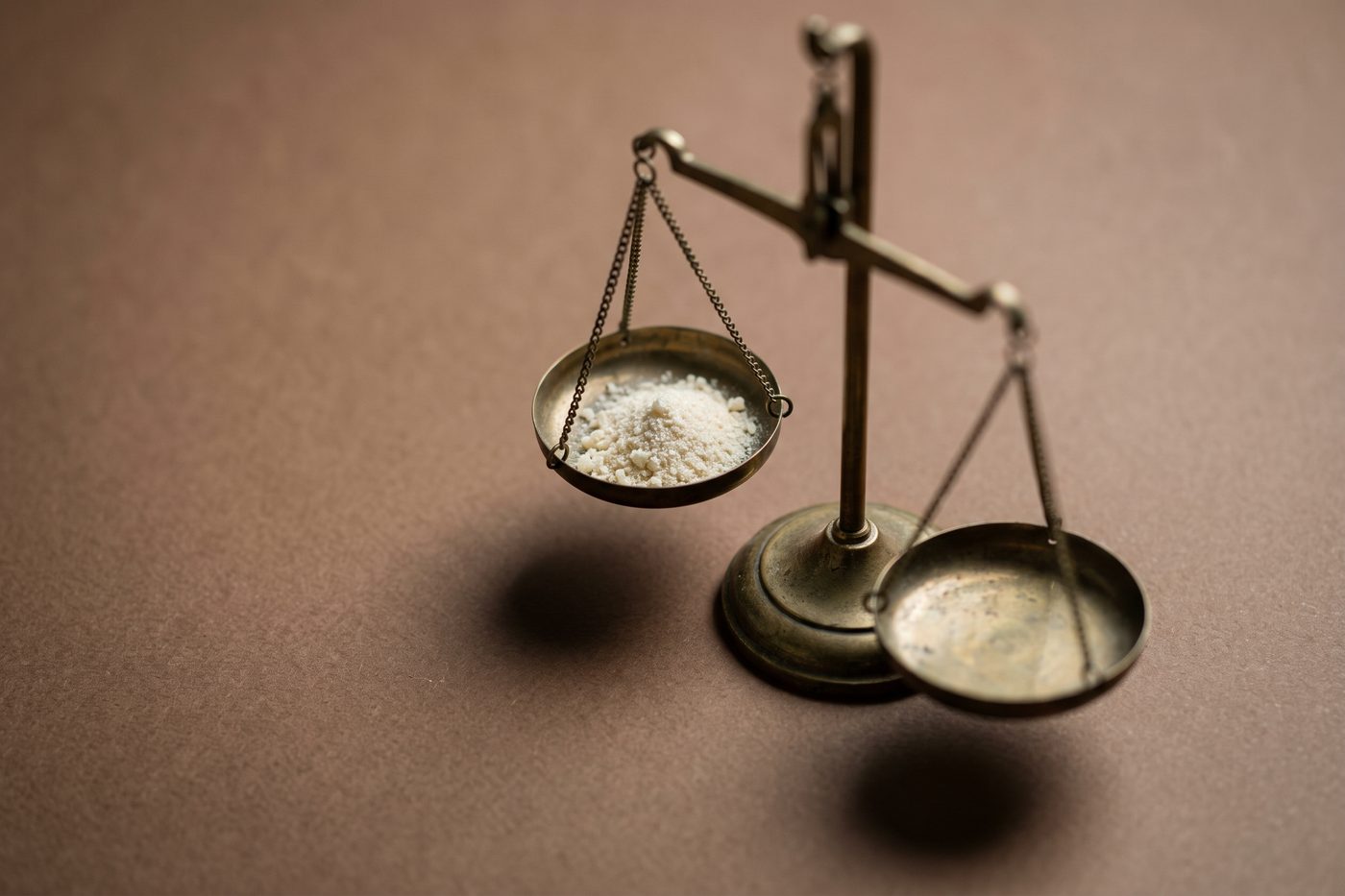 An antique brass balance scale, one shallow pan holding a small heap of pale crystalline powder, matte navy backdrop