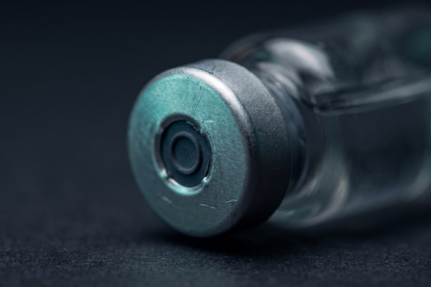 A crimped aluminium seal and grey rubber stopper, the glass beneath softly out of focus, cool teal rim light on navy