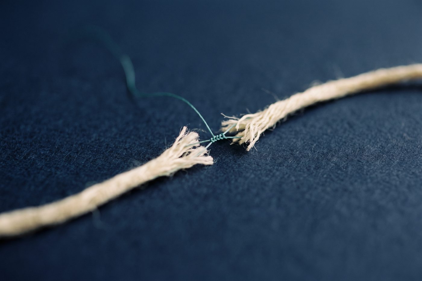 A frayed natural linen fibre bridged and knitted back together by a single fine teal filament on matte navy, extreme macro
