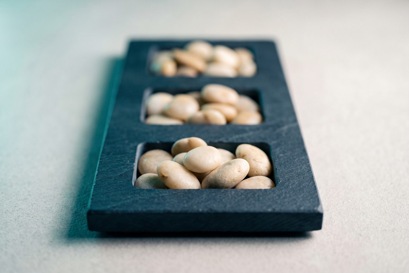 A navy slate slab carved with three recesses, all three filled with smooth pale stones, raking teal light