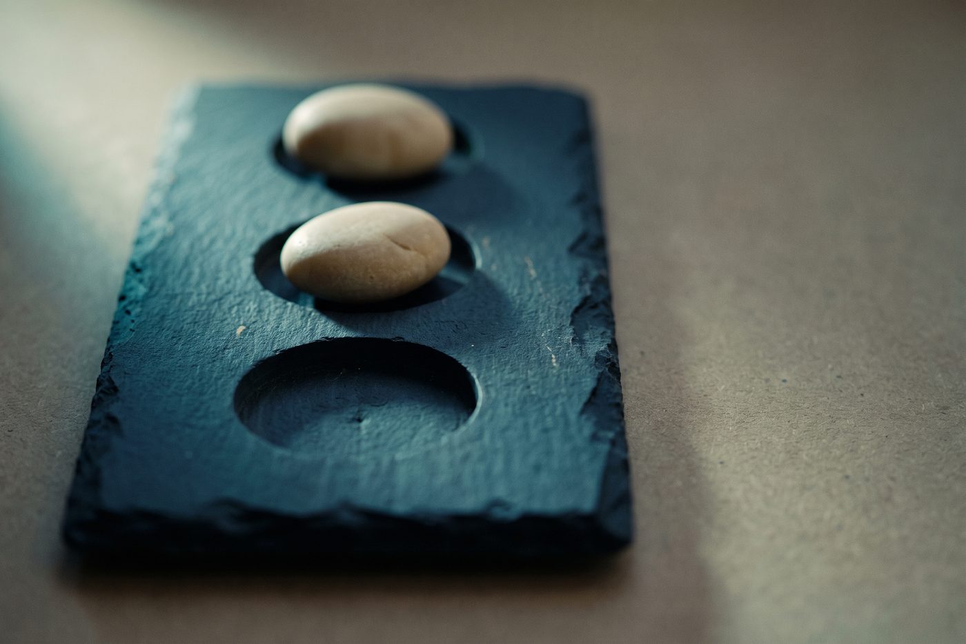 A navy slate slab carved with three recesses, two smooth pale stones seated in them and one left empty, raking teal light