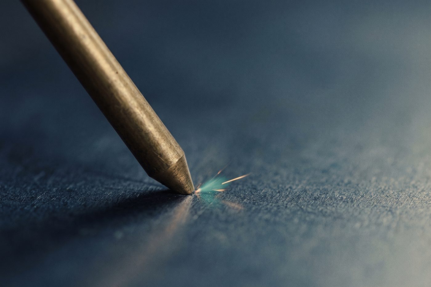 A fine brass pin striking one precise point on a steel plate, a single teal spark, extreme macro, navy