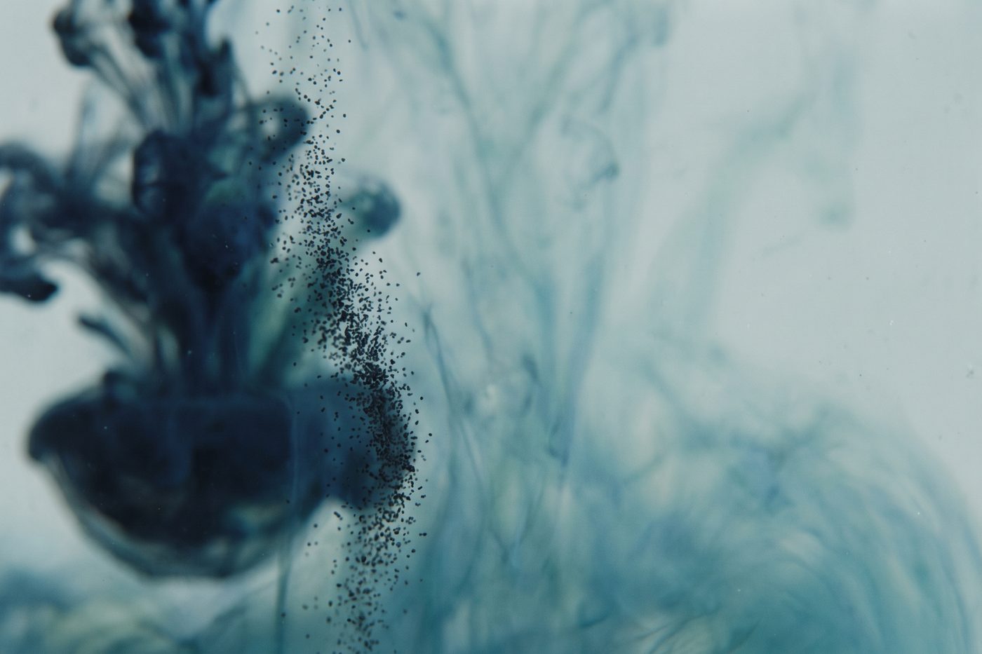 Fine dark pigment granules dispersing into clear water, strongly backlit, navy and teal