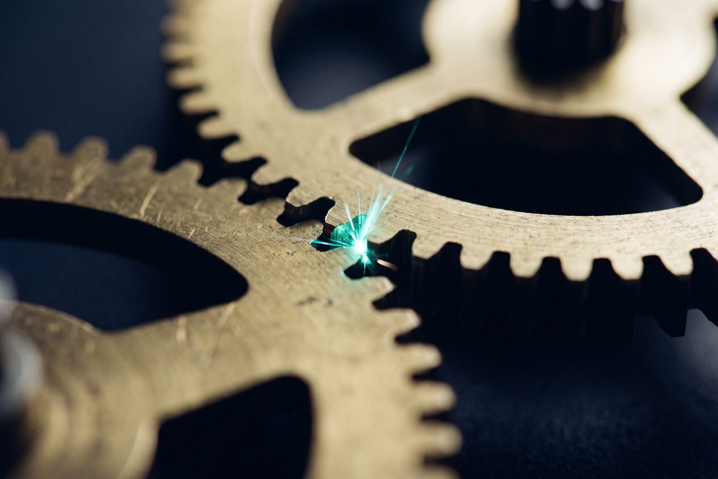 Brass cogs meshing in extreme macro with a bright teal spark at the point of contact, deep navy shadow