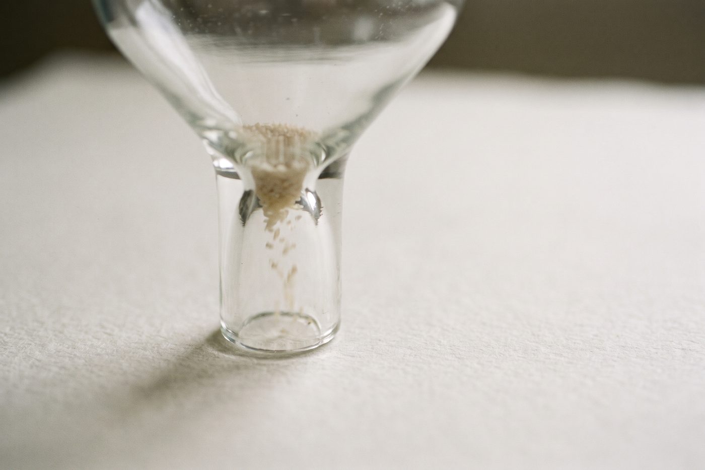 A sealed glass funnel narrowing to a thin neck, a few pale grains passing through the constriction, backlit teal on navy