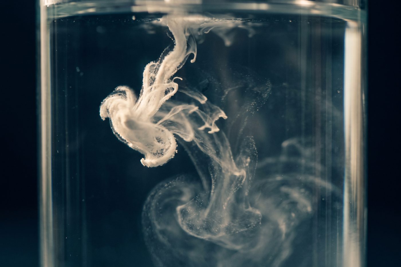 A trail of white powder unfurling and dissolving through clear water inside a glass vessel, strongly backlit, cool teal and navy