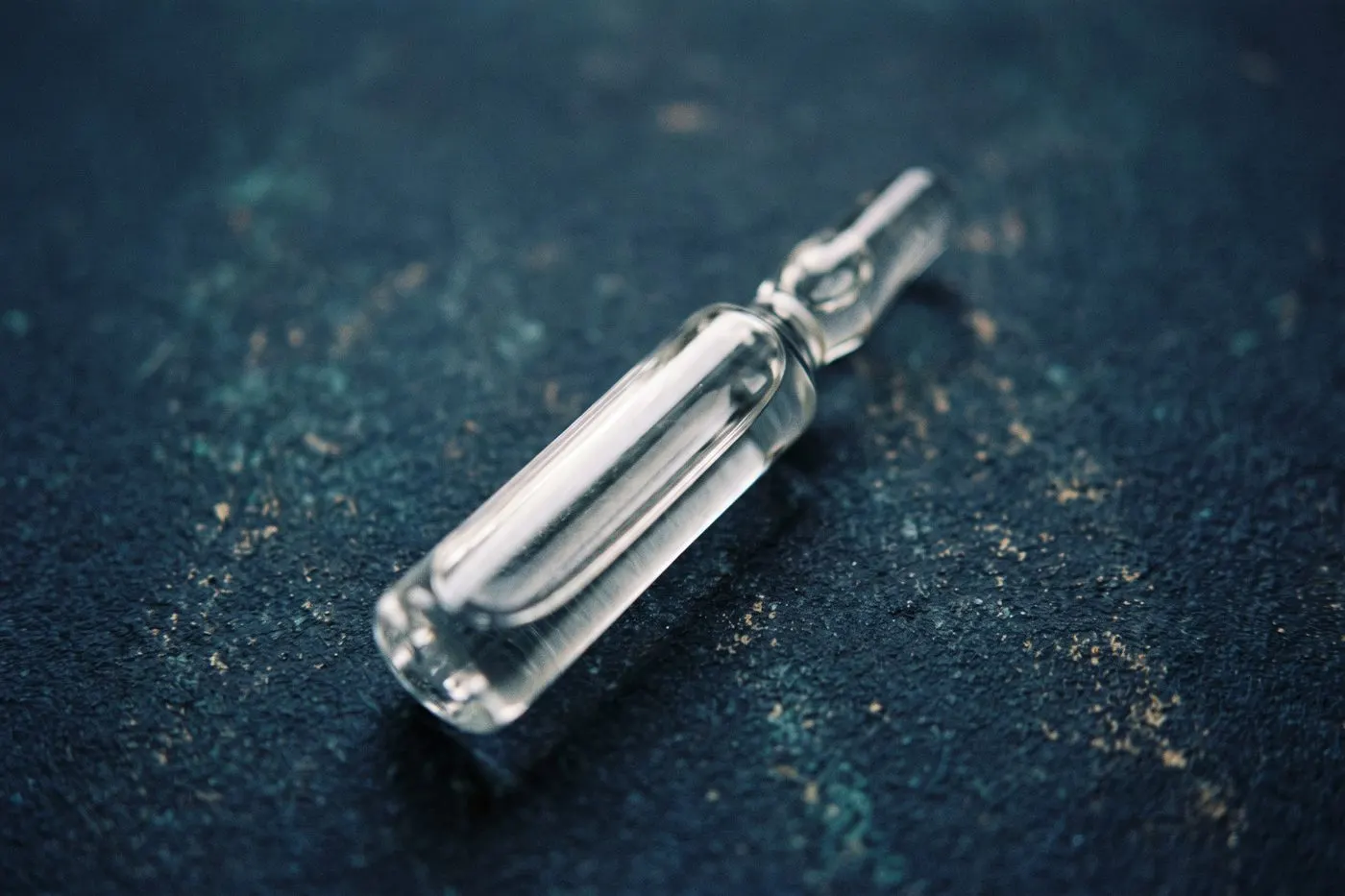 A sealed glass ampoule of clear liquid lying on matte navy stone, a single long highlight running its length