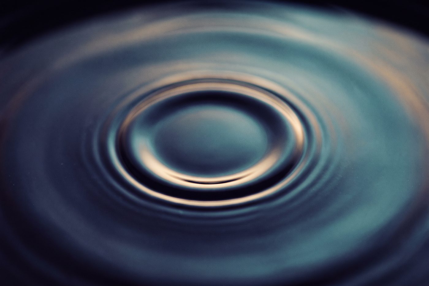 A still pool of dark water with one perfect expanding ring at its centre, cool teal light