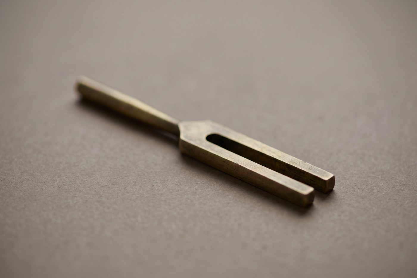 A brass tuning fork with an elongated handle resting on navy slate, warm cream raking light