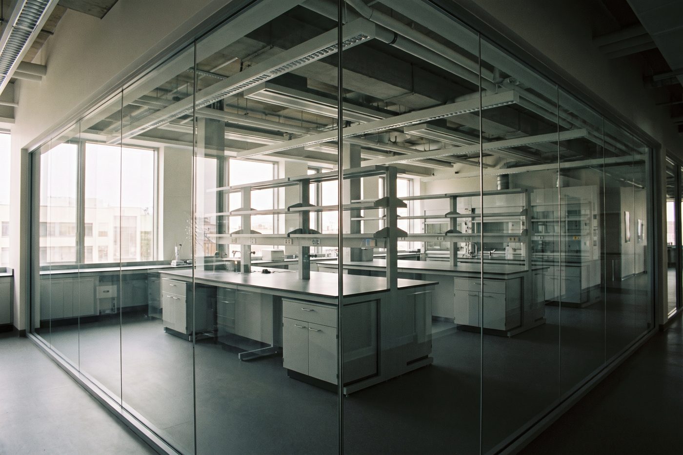 A modern laboratory interior seen through a glass partition, stainless benches and cool teal light, completely deserted