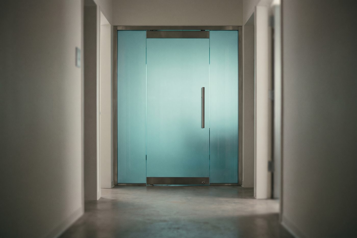A frosted glass door panel in a quiet laboratory corridor, teal light glowing behind it, a brushed steel handle in sharp focus