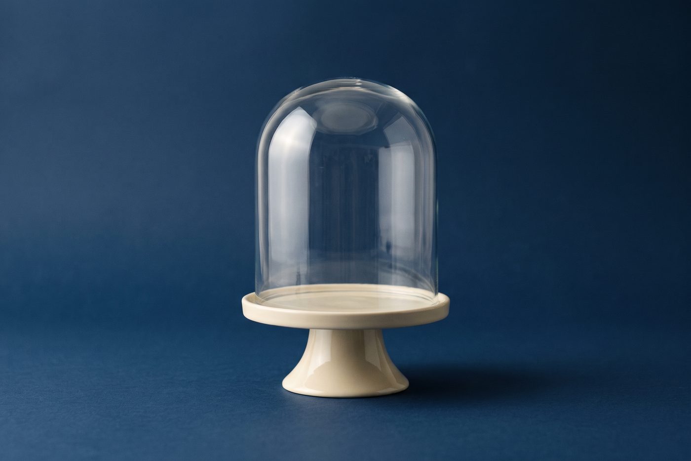 A sealed glass bell jar over an empty cream pedestal on a navy surface, cool light, strong negative space