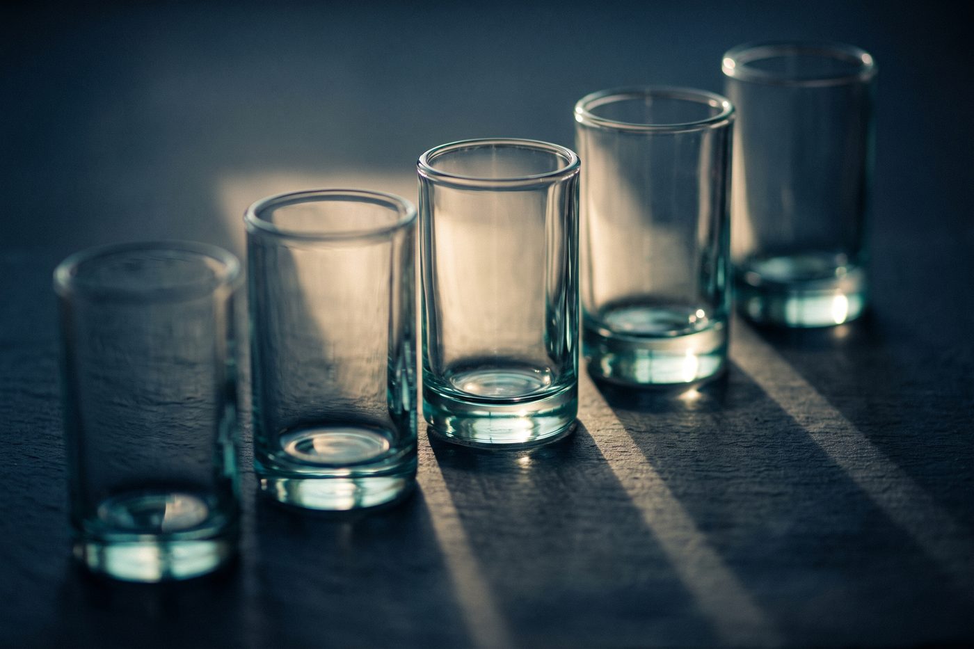 A row of five identical glass cylinders of ascending height on navy slate, raking cream light throwing stepped shadows